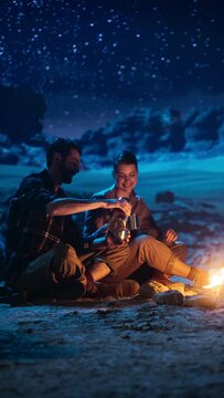 Vertical Video: Happy Couple Sitting by Campfire Sharing Cup of Drink, Watching Night Sky while Camping in the Canyon. Two Traveling people Tell Inspirational Stories, Look at Milky Way Stars