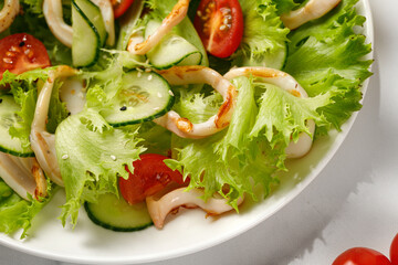 Grilled squid rings or calamari on green salad leaves, with tomatoes, cucumber on white plate.  Close up