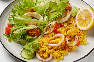 Dish with squid rings, rice, green vegetable salad with tomatoes, cucumber on white plate.