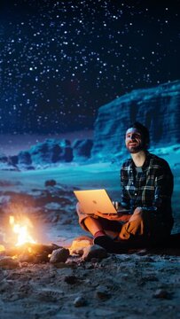Vertical Video: Male Traveler Sitting by Campfire Uses Laptop Computer while Tent Camping in the Canyon. Man doing Remote Work, e-business, e-shopping, ecommerce, using Social Media Internet