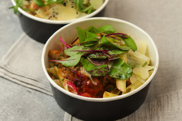 Pasta with mussels, cream sauce, parmesan and greens in a round paper takeaway food container on napkin on a gray background. Concept of take-away and healthy food delivery. Close-up, top view.