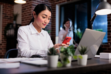 Businesswoman at work sending messages on social media platform using mobile phone. Happy smiling asian office worker sitting in office workspace while texting somebody on modern smartphone device.