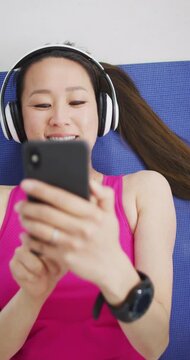 Vertical video of smiling asian woman relaxing wearing headphones and using smartphone