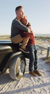 Vertical video of caucasian gay male couple sitting on car at beach and embracing