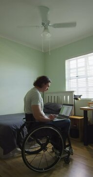Vertical video of caucasian disabled man sitting in wheelchair using laptop