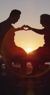 Vertical video of caucasian gay male couple sitting on car at sunset beach making heart with hands