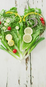 Vertical video of fresh tomatoes and green vegetables forming heart on white rustic background