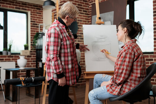 Senior art teacher showing graphic technique to young student during creativity lesson in school studio. Painter woman drawing vase sketch using sketching pencil working at personal growth