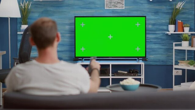 Man sitting on sofa looking at green screen on tv and switching channels in modern living room. Back view of person relaxing on couch using television remote zapping programs on chroma key display.