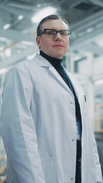 Vertical Portrait of a Professional Male Heavy Industry Engineer/Worker Wearing White Laboratory Robe and Safety Glasses. Confident Caucasian Industrial Specialist Standing in a Factory Facility.