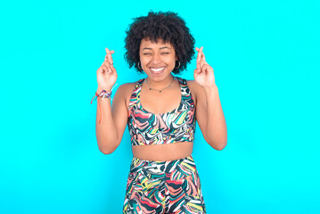 Joyful young woman with afro hairstyle in sportswear against blue background clenches teeth, raises fingers crossed, makes desirable wish, waits for good news, I have to win.