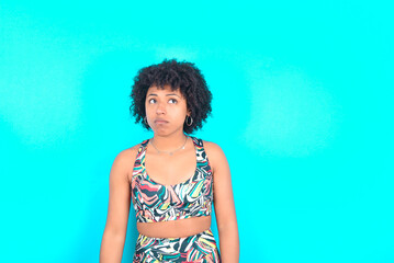 young woman with afro hairstyle in sportswear against blue background has worried face looking up lips together, being upset thinking about something important, keeps hands down.