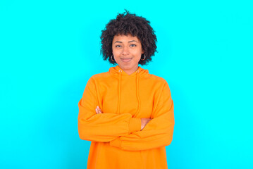 Obraz na płótnie Canvas Self confident serious calm young woman with afro hairstyle wearing orange hoodie against blue background stands with arms folded. Shows professional vibe stands in assertive pose.