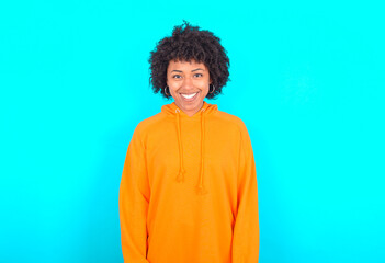 young woman with afro hairstyle wearing orange hoodie against blue background with nice beaming smile pleased expression. Positive emotions concept