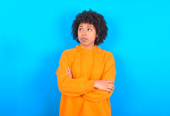 Obraz na płótnie Canvas Charming thoughtful young woman with afro hairstyle wearing orange hoodie against blue background stands with arms folded concentrated somewhere with pensive expression thinks what to do