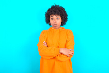 Obraz na płótnie Canvas Shocked embarrassed young woman with afro hairstyle wearing orange hoodie against blue background keeps mouth widely opened. Hears unbelievable novelty stares in stupor