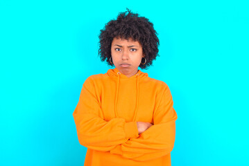 Obraz na płótnie Canvas Gloomy dissatisfied young woman with afro hairstyle wearing orange hoodie against blue background looks with miserable expression at camera from under forehead, makes unhappy grimace