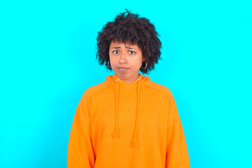 Obraz na płótnie Canvas Offended dissatisfied young woman with afro hairstyle wearing orange hoodie against blue background with moody displeased expression at camera being disappointed by something