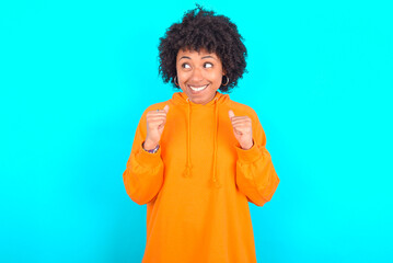 Obraz na płótnie Canvas young woman with afro hairstyle wearing orange hoodie against blue background clenches fists and awaits for something nice happened looks away bites lips and waits announcement of results