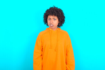 Obraz na płótnie Canvas Portrait of dissatisfied young woman with afro hairstyle wearing orange hoodie against blue wall smirks face, purses lips and looks with annoyance at camera, discontent hearing something unpleasant