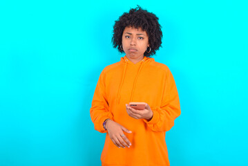 Obraz na płótnie Canvas Upset dissatisfied young woman with afro hairstyle wearing orange hoodie against blue background uses mobile software application and surfs information in internet, holds modern mobile hand