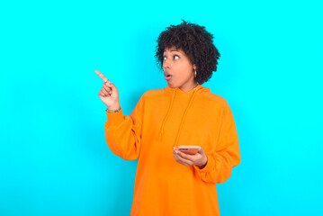 Stunned young woman with afro hairstyle wearing orange hoodie against blue background points sideways right copy space, recommends product, sees astonishing thing