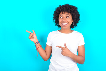 Optimistic young woman with afro hairstyle wearing white T-shirt against blue wall points with both hands and  looking at empty space.