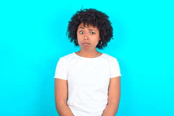 Displeased young woman with afro hairstyle wearing white T-shirt against blue wall frowns face feels unhappy has some problems. Negative emotions and feelings concept