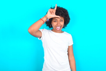 Funny young woman with afro hairstyle wearing white T-shirt against blue wall makes loser gesture mocking at someone sticks out tongue making grimace face.