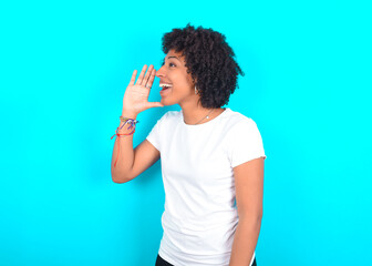 young woman with afro hairstyle wearing white T-shirt against blue wall look empty space holding hand near her face and screaming or calling someone.