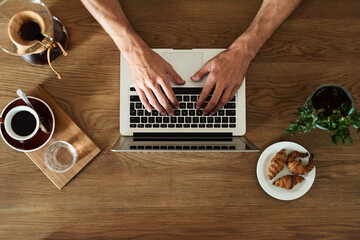 Man's hands working on laptop at breakfast. Top view of unrecognizable businessman working on laptop at table with coffee cup, chemex, plate of croissants and plant.