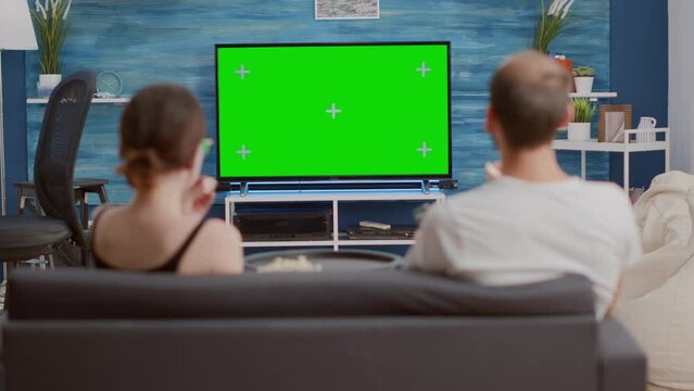 Static tripod shot of couple having a snack while looking at green screen tv watching a movie and turning up the volume. Woman and man watching chroma key television and zapping while sharing popcorn.