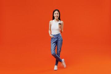 Full size smiling excited young woman of Asian ethnicity 20s years old in white tank top hold takeaway delivery craft paper brown cup coffee to go isolated on plain orange background studio portrait.