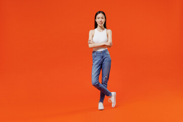 Full size body length smiling young woman of Asian ethnicity 20s years old in white tank top hold hands crossed isolated on plain orange background studio portrait. People emotions lifestyle concept.