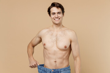 Fototapeta premium Satisfied shirtless young man 20s with naked torso showing loose pants isolated on pastel pastel beige color background Body care healthcare fitness sport bodybuilding diet loosing weight concept.