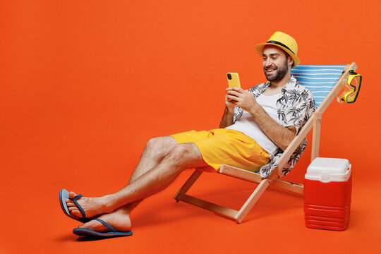 Young fun happy tourist man in beach shirt hat lie on deckchair near fridge hold use mobile cell phone isolated on plain orange background studio portrait. Summer vacation sea rest sun tan concept