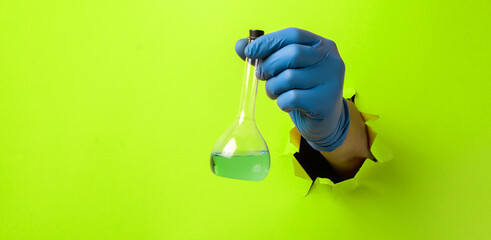 The glass flask with green liquid in hand through a torn hole in the green paper.