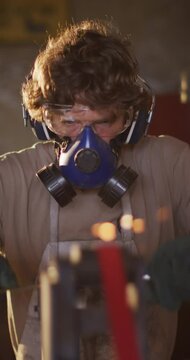 Vertical video of caucasian male blacksmith in mask and goggles grinding metal tool in workshop