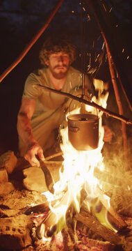 Vertical video of bearded caucasian male survivalist cooking over campfire in wilderness