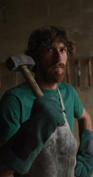 Vertical video portrait of bearded caucasian male blacksmith holding hammer in workshop