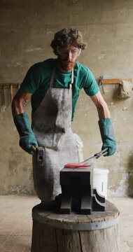Vertical video of caucasian male blacksmith forging hot metal with hammer on anvil in workshop