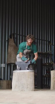 Vertical video of caucasian male blacksmith forging hot metal with hammer on anvil in workshop