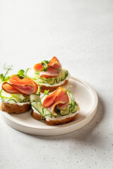 Bruschetta with baguette, bacon or meat, cream cheese, micro-greenery, fresh cucumber and sprouts, on white plate on white textured background