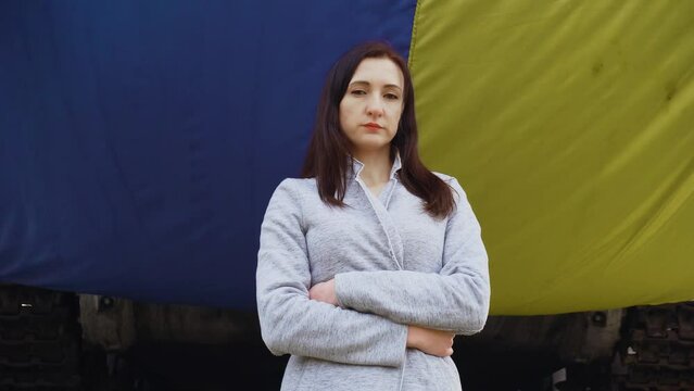 Serious refugee woman standing near big waving Ukrainian flag in foreign country during invasion. Crisis, peace, stop aggression in whole world