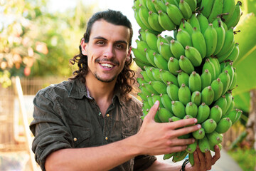 A young hipster farmer looks admiringly and holding bananas at an organic farm with a smile and happiness cause ready for harvest. Labor, hard work, hope, and a sustainable living of concept ideas © tai