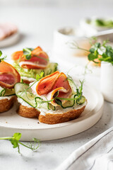 Three Bruschettas with baguette, bacon or meat, cream cheese, micro-greenery, fresh cucumber and sprouts, in composition on white plate on white textured background