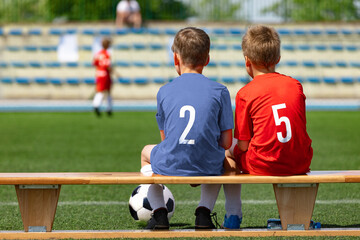 Two boys' sports friends sitting on a wooden bench and watching a soccer match. School kids in football uniforms. Happy children playing sports outdoor. Friendship between two opposite sports teams - Powered by Adobe