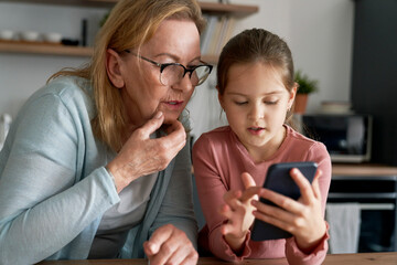 Caucasian grandmother and her granddaughter using smart phone together and sitting in the kitchen
