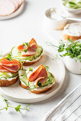 Bruschettas with baguette, bacon or meat, cream cheese, micro-greenery, fresh cucumber and sprouts, in composition on white plate on white textured background