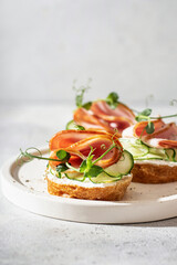 Three bruschettas with baguette, bacon or meat, cream cheese, micro-greenery, fresh cucumber and sprouts, on white plate on white textured background